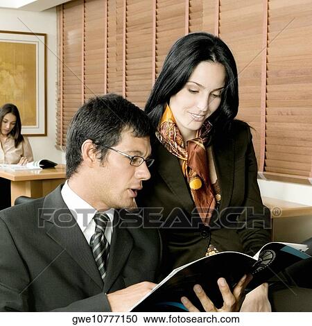 Stock Image - Businessman and a businesswoman sitting in an office and looking at a magazine. Fotosearch