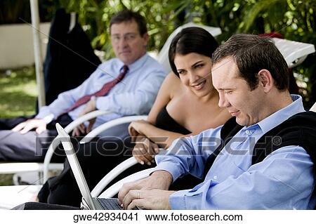 Businessman and a businesswoman working on a laptop with another businessman sitting beside them View Large Photo Image Stock Image - Businessman and a businesswoman working on a laptop with another businessman sitting beside them. Fotosearch