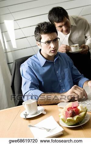 Businessman using a laptop with another businessman standing behind him View Large Photo Image Stock Image - Businessman using a laptop with another businessman standing behind him. Fotosearch