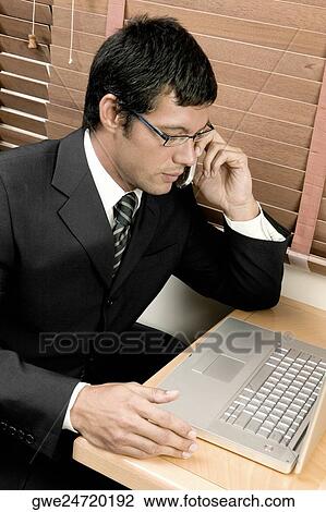 Stock Image - High angle view of a businessman sitting in front of a laptop and talking on a mobile phone. Fotosearch