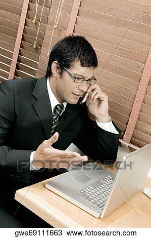 Stock Image - High angle view of a businessman sitting in front of a laptop using a mobile phone. Fotosearch