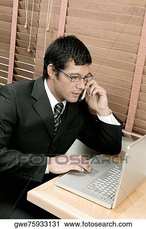 Stock Image - High angle view of a businessman sitting in front of a laptop using a laptop and talking on a mobile phone. Fotosearch