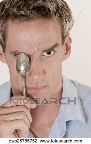 Stock Image - Portrait of a mid adult man covering his eye with a spoon. Fotosearch