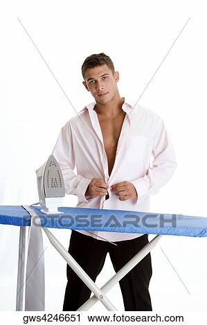 Stock Image - Portrait of a young man standing in front of an ironing board and buttoning his shirt. Fotosearch