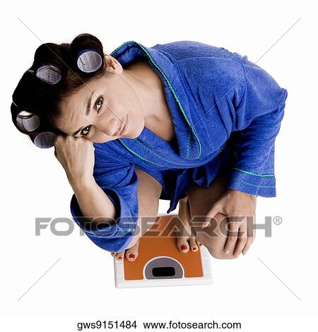 Picture - Portrait of a young woman with curlers in her hair standing on a weight scale. Fotosearch