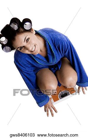 Stock Image - Portrait of a young woman with curlers in her hair crouching on a weight scale. Fotosearch
