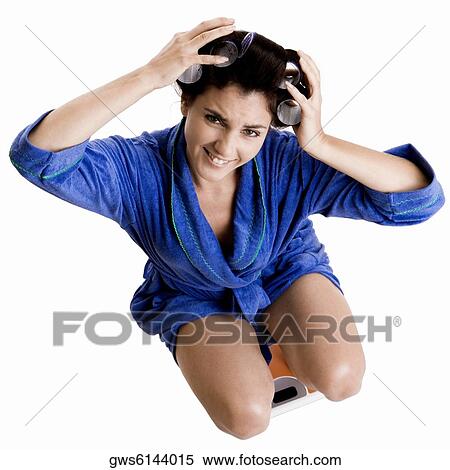 Stock Photography - Portrait of a young woman with curlers in her hair crouching on a weight scale. Fotosearch