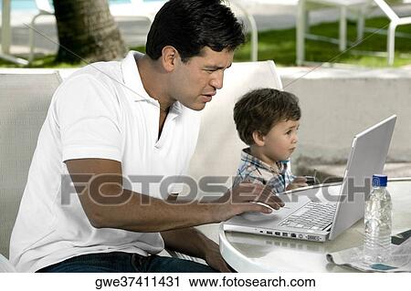 Stock Image - Side profile of a father working on a laptop with his son sitting beside him. Fotosearch