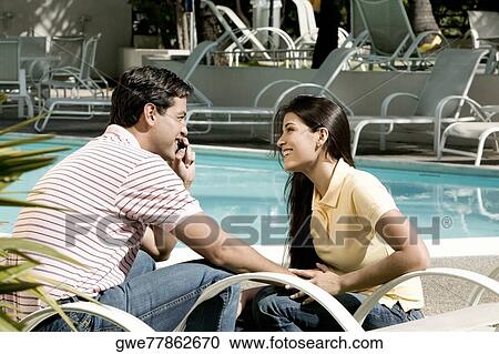 Stock Image - Side profile of a young man talking on a mobile phone with a young woman sitting in front of him. Fotosearch