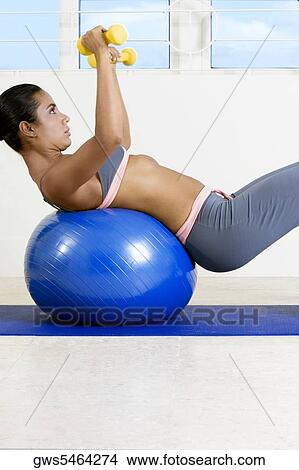Side profile of a young woman exercising with dumbbells on a fitness ball View Large Photo Image Picture - Side profile of a young woman exercising with dumbbells on a fitness ball. Fotosearch