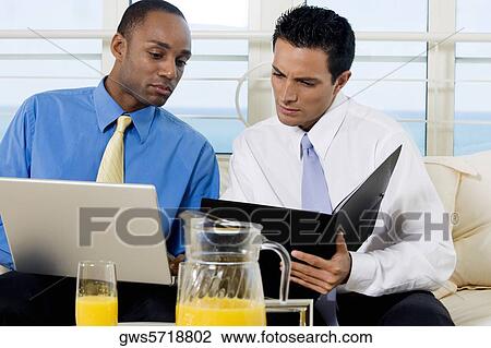 Two businessmen sitting on a couch holding a file and a laptop View Large Photo Image Stock Image - Two businessmen sitting on a couch holding a file and a laptop. Fotosearch