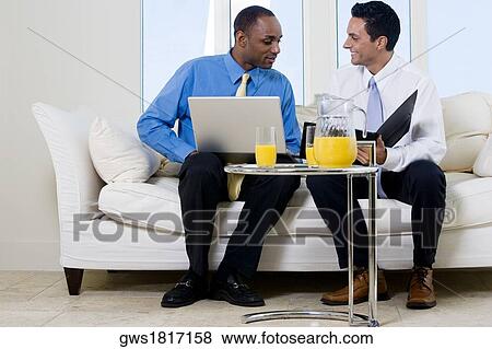 Two businessmen sitting on a couch holding a file and a laptop View Large Photo Image Stock Photo - Two businessmen sitting on a couch holding a file and a laptop. Fotosearch