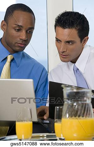 Two businessmen sitting on a couch looking at a laptop View Large Photo Image Stock Photo - Two businessmen sitting on a couch looking at a laptop. Fotosearch