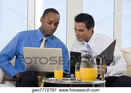 Two businessmen sitting on a couch looking at a laptop View Large Photo Image Stock Photo - Two businessmen sitting on a couch looking at a laptop. Fotosearch