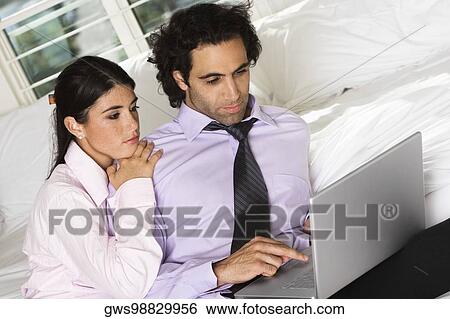 Stock Photograph - Close-up of a businessman and a businesswoman sitting on the bed using a laptop. Fotosearch