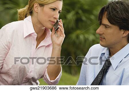 Stock Photograph - Close-up of a businesswoman talking on a mobile phone with a businessman looking at her. Fotosearch