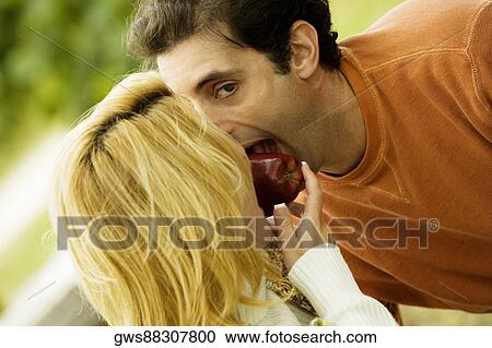 Stock Image - Close-up of a young woman and a mid adult man biting an apple together. Fotosearch