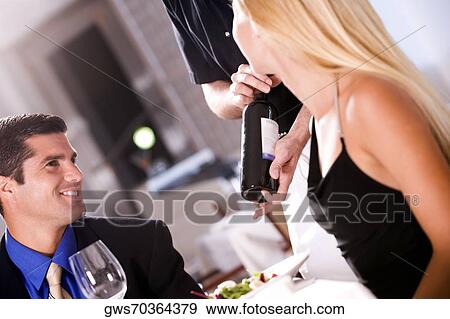 Stock Photo - Mid adult man and a young woman sitting at the table with a waiter showing them a wine bottle. Fotosearch