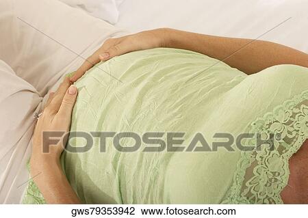 Stock Image - Mid section view of a pregnant woman lying on the bed touching her abdomen. Fotosearch