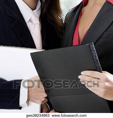 Stock Image - Mid section view of two businesswomen holding a file and a laptop. Fotosearch