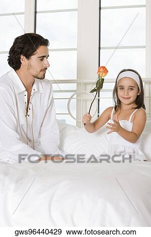 Stock Photo - Portrait of a girl sitting and holding a rose with her father sitting beside her. Fotosearch