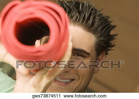 Stock Image - Portrait of a young man looking through a rolled up exercise mat. Fotosearch