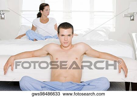 Stock Image - Portrait of a young man sitting on the floor with a young woman sitting behind him on the bed. Fotosearch