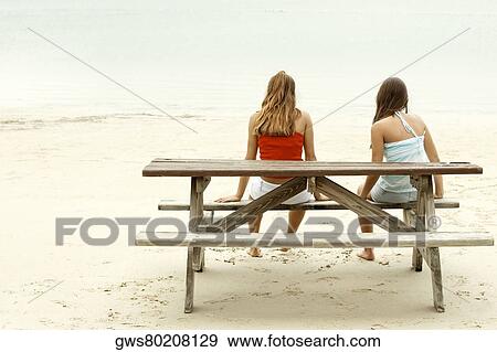Stock Photo - Rear view of a girl and a teenage girl sitting on a picnic table on the beach. Fotosearch