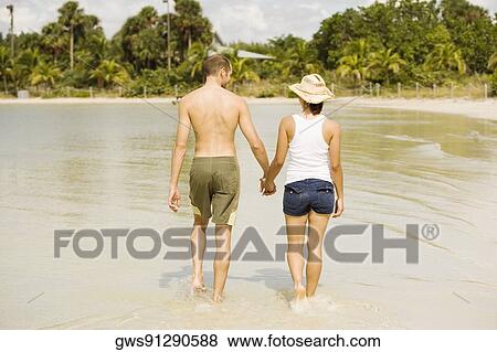 Stock Photo - Rear view of a young man and a teenage girl holding hands walking on the beach. Fotosearch