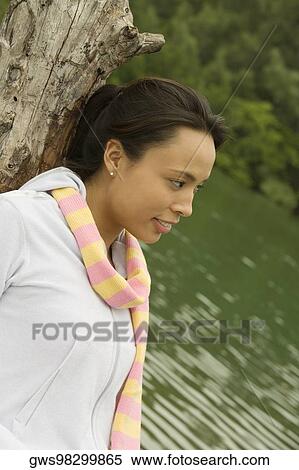 Stock Photography - Side profile of a young woman leaning against a tree trunk by the lake. Fotosearch