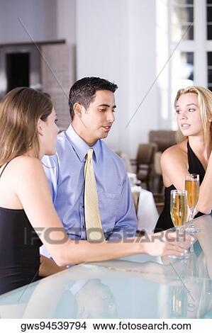 Picture - Two young women and a mid adult man sitting at a bar counter. Fotosearch