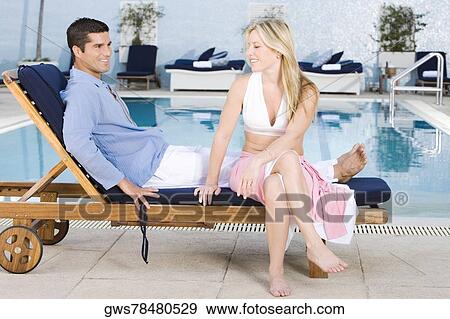 Stock Photo - Young woman and a mid adult man sitting on a deck chair at the poolside. Fotosearch