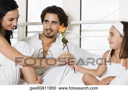 Young woman sitting on the bed giving a flower to her to her daughter with a young man sitting beside her View Large Photo Image Stock Image - Young woman sitting on the bed giving a flower to her to her daughter with a young man sitting beside her. Fotosearch