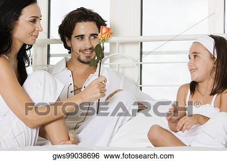 Young woman sitting on the bed holding a flower with a young man and her daughter View Large Photo Image Stock Photograph - Young woman sitting on the bed holding a flower with a young man and her daughter. Fotosearch