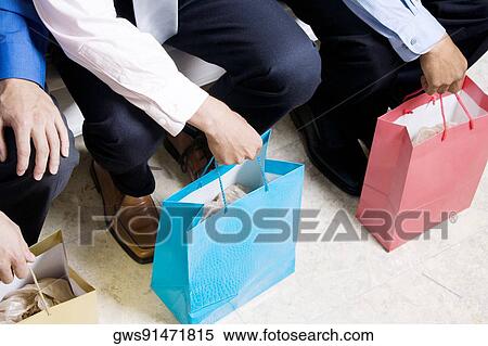 Stock Photography - Low section view of three businessmen sitting with shopping bags. Fotosearch