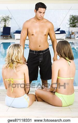Stock Image - Mid adult man standing in front of two young women at the poolside. Fotosearch