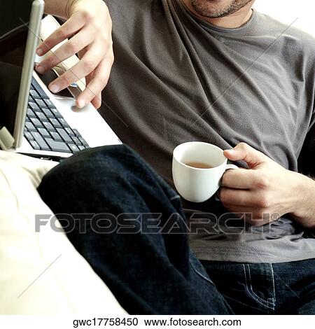 Stock Image - Mid section view of a man holding a cup of tea using a laptop. Fotosearch