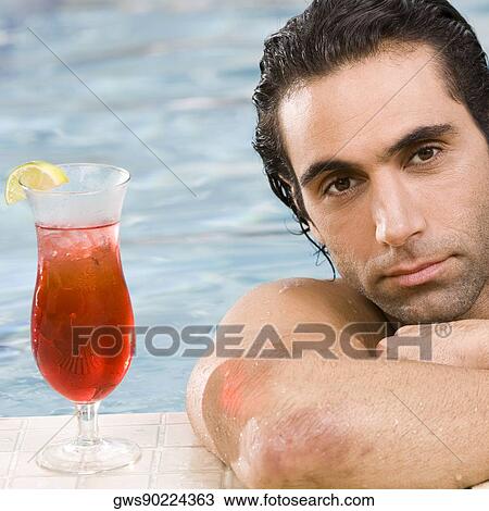 Stock Image - Portrait of a mid adult man leaning at the edge of a swimming pool with a cocktail. Fotosearch
