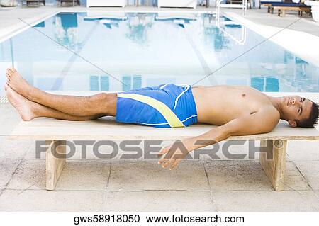 Stock Image - Portrait of a young man lying down on a bench at the poolside. Fotosearch