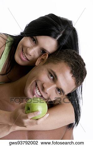 Stock Image - Portrait of a young woman holding a green apple embracing a young man from behind. Fotosearch