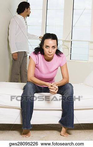 Portrait of a young woman sitting on the bed with a young man standing behind her View Large Photo Image Stock Photograph - Portrait of a young woman sitting on the bed with a young man standing behind her. Fotosearch