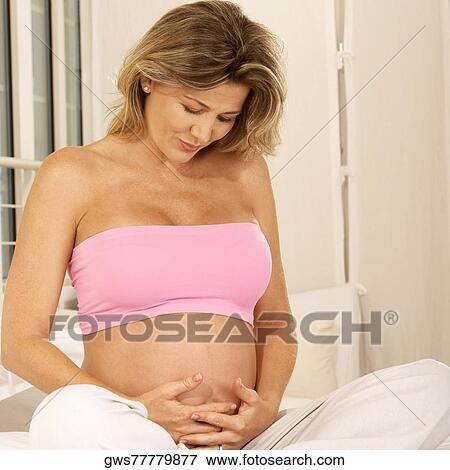 Pregnant woman sitting on the bed touching her abdomen View Large Photo Image Stock Photo - Pregnant woman sitting on the bed touching her abdomen. Fotosearch