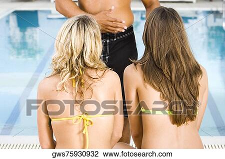 Stock Image - Rear view of two young women listening to a mid adult man at the poolside. Fotosearch