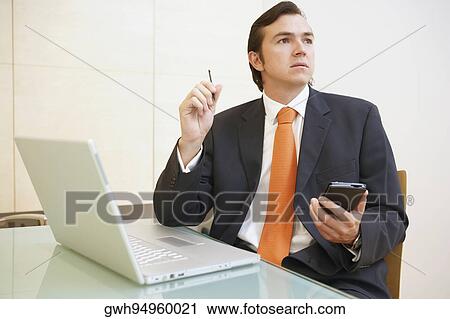 Stock Image - Close-up of a businessman holding a personal data assistant in front of a laptop. Fotosearch