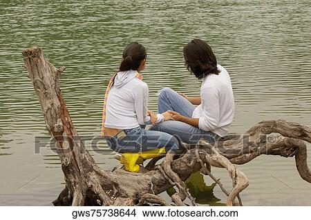 High angle view of a young couple sitting on a tree over a lake View Large Photo Image Picture - High angle view of a young couple sitting on a tree over a lake. Fotosearch