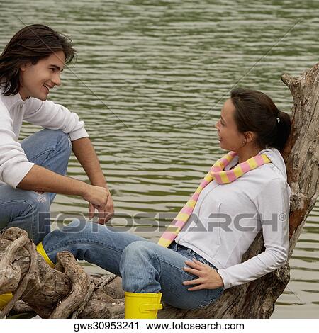 Stock Image - High angle view of a young couple sitting on a tree over a lake. Fotosearch