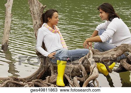 Stock Image - High angle view of a young couple sitting on a tree over a lake. Fotosearch
