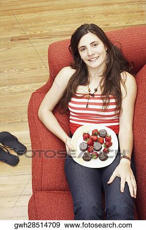 Stock Photo - High angle view of a young woman lying on a couch and holding a plate of strawberries with chocolates. Fotosearch