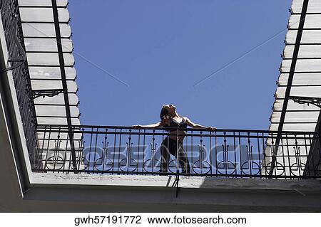 Stock Image - Low angle view of a young woman standing in the balcony of a building . Fotosearch