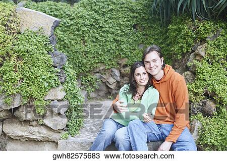 Stock Image - Portrait of a young woman and a mid adult man sitting on a rock and embracing each other. Fotosearch